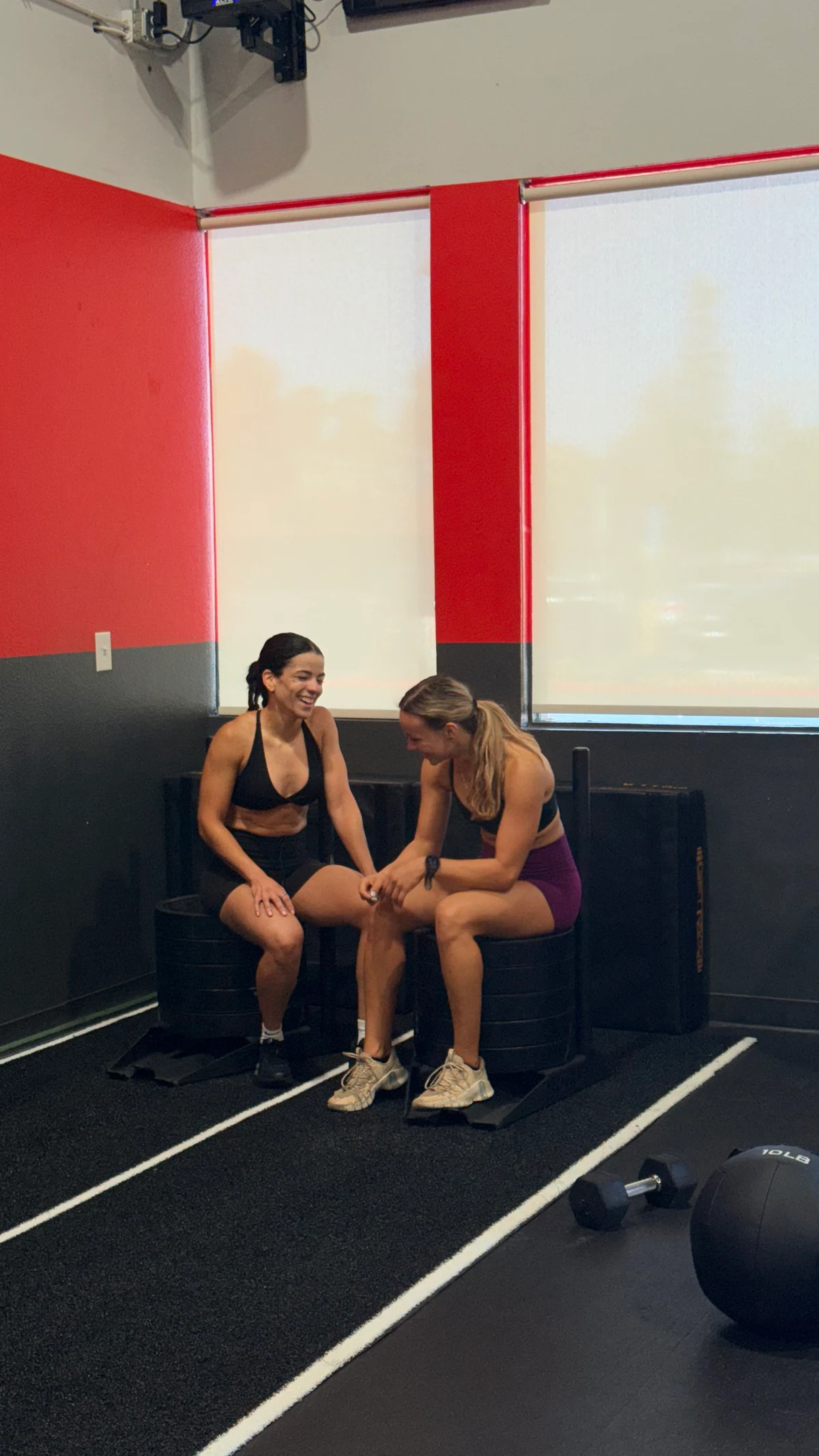 Two women laughing while sitting on a loaded push sled on a turf lane in a gym with red and black walls
