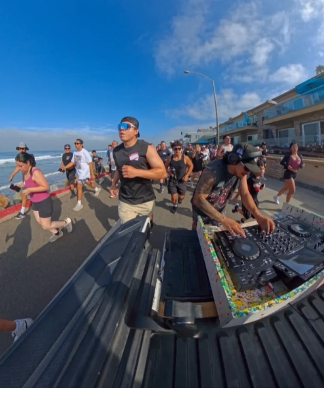 Runners spread across the Oceanside beachfront road on a bright morning, with a DJ playing off the open tailgate of a truck ahead of them