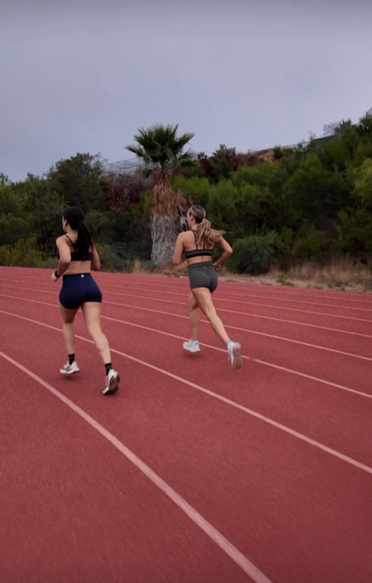 Two women running away from the camera down a red track, with a palm tree and green hillside ahead under a gray sky