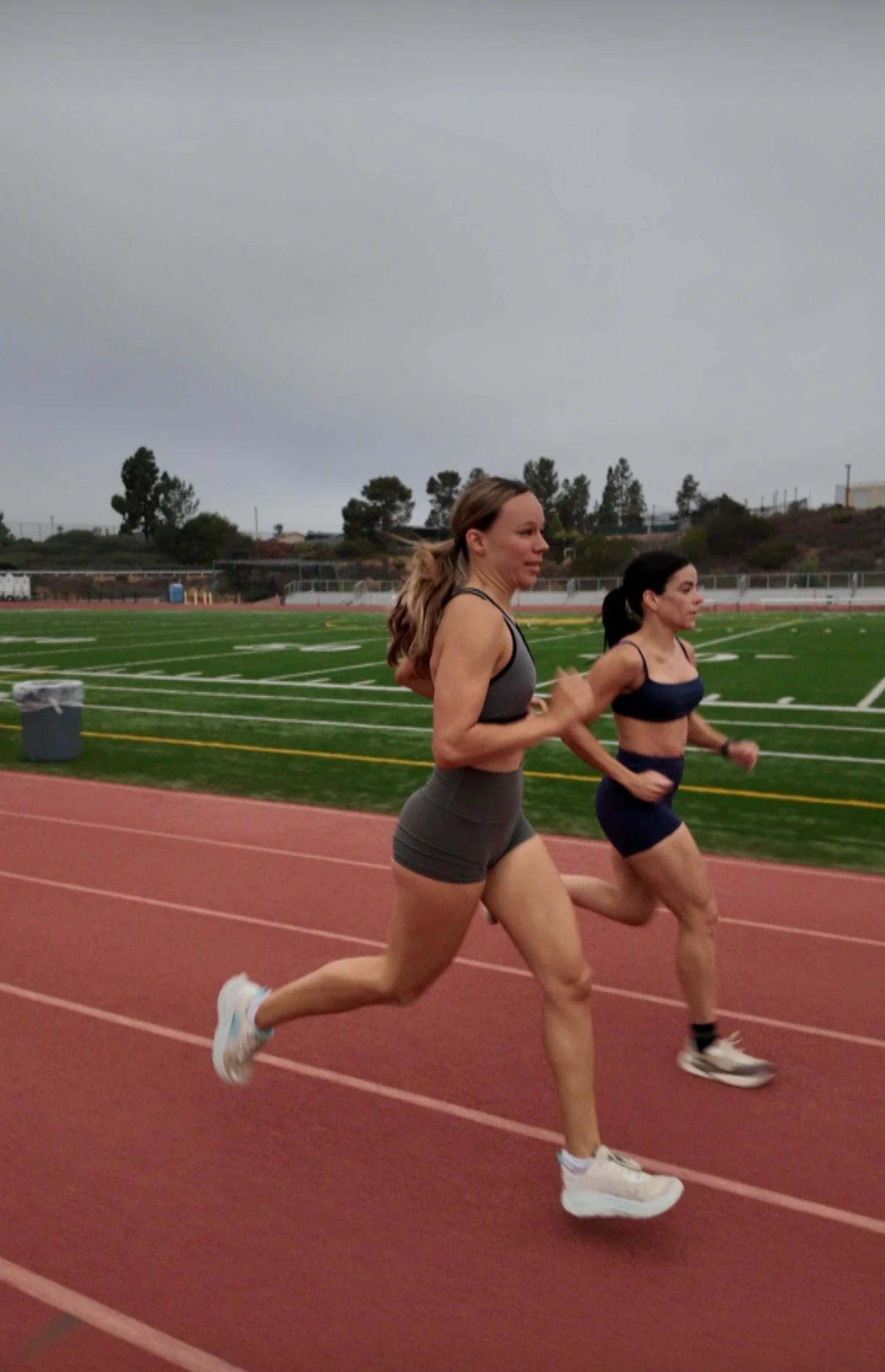Two women running side by side on a red track next to a football field under an overcast sky